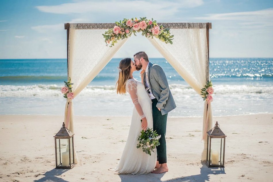 Barefoot Beach Arch for Intimate Vows Bride and groom kissing under a rustic floral beach arch with lanterns on the sand beside the ocean.