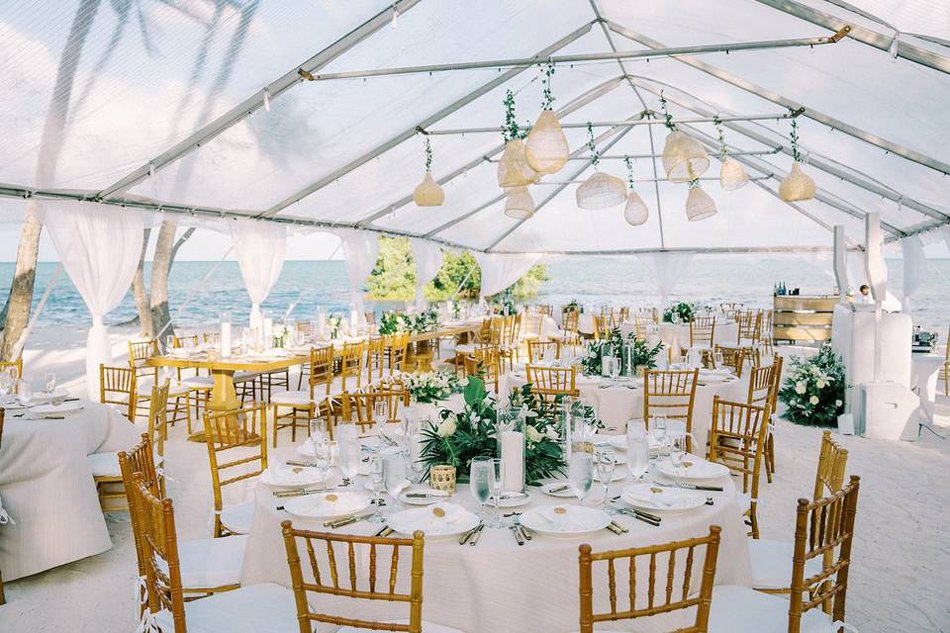 Clear-Top Tent Reception on the Sand Coastal reception under a clear tent with woven pendants, round tables, and ocean backdrop on the beach.