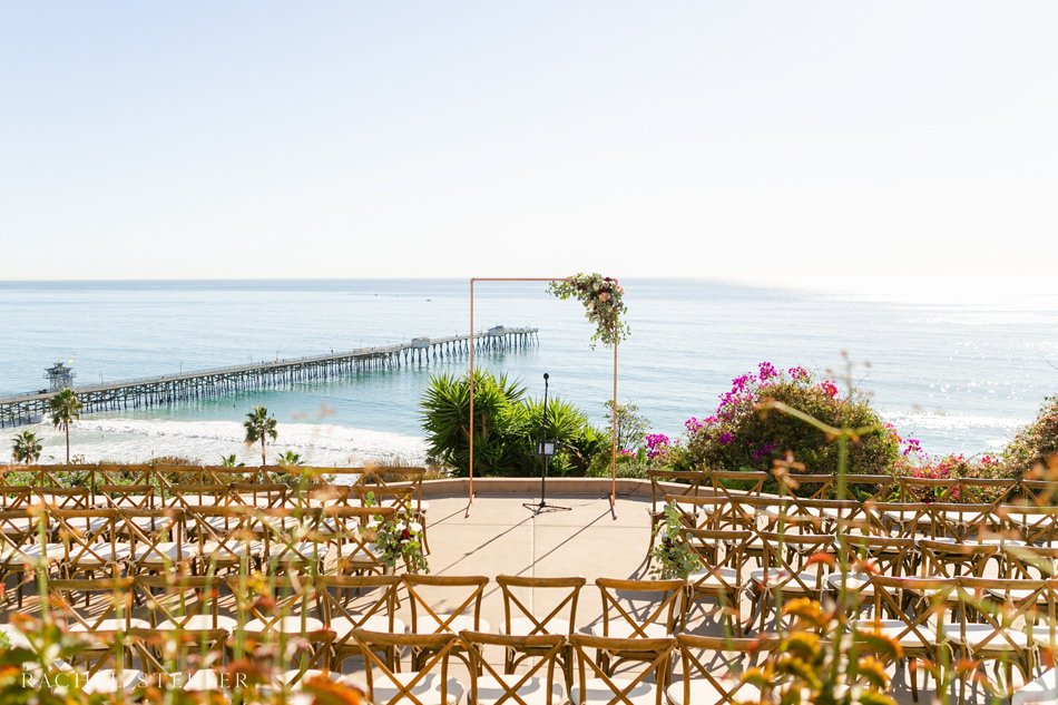 Oceanview Terrace with Pier Backdrop Clifftop terrace ceremony overlooking the Pacific with a coastal pier in the distance and seasonal blooms.