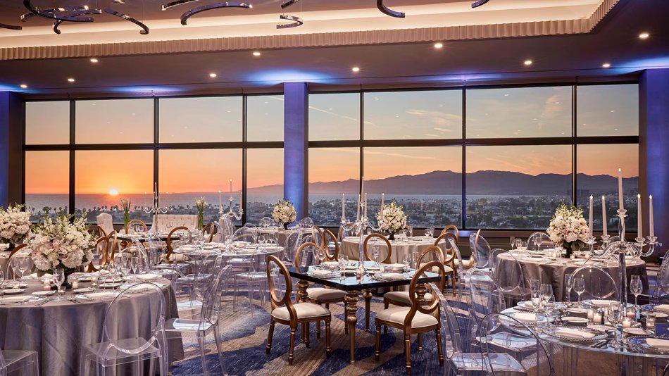 Marina del Rey Sunset Ballroom Glass-walled hotel ballroom at sunset overlooking Marina del Rey with round tables and ghost chairs.