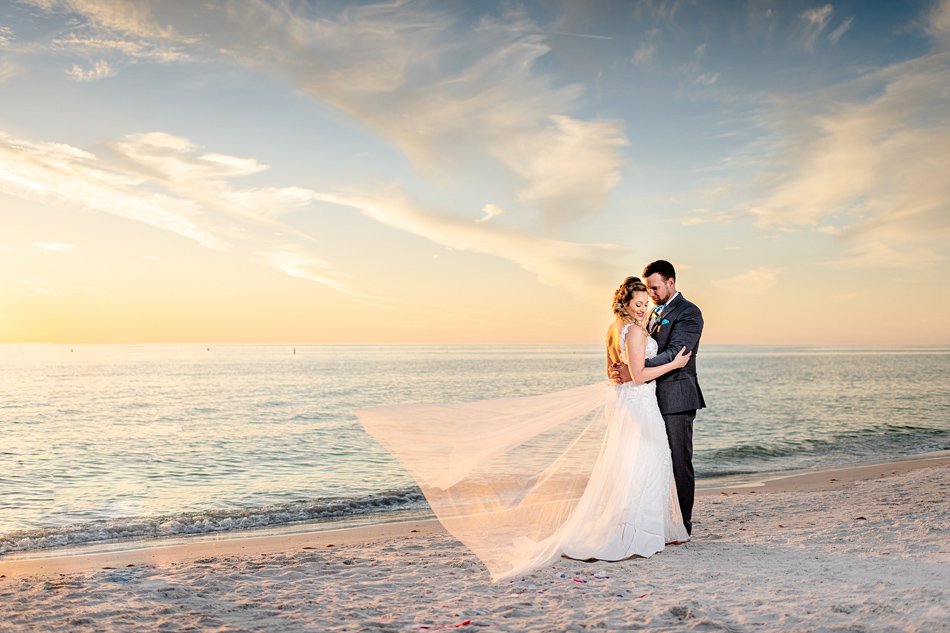 Couple embraces on a Southern California beach at sunset with flowing veil and gentle waves—a refined luxury wedding scene.
