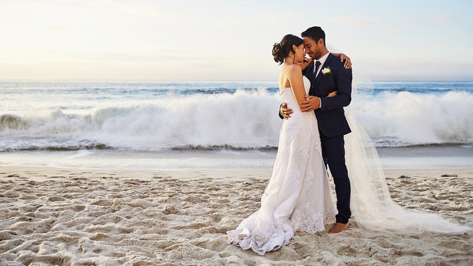 Bride and groom embrace on a Southern California beach at sunset with waves rolling behind them.
