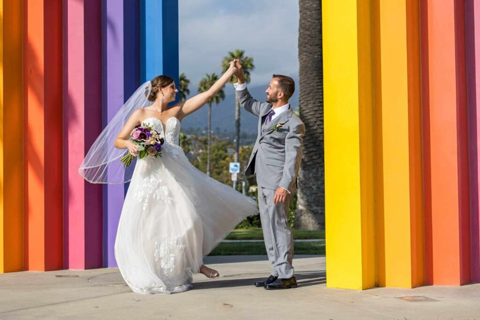 Bride and groom twirl at Santa Barbara’s Chromatic Gate near East Beach—colorful columns, palm trees, and Pacific coastline in Southern California.