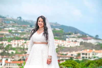 Elegant South Asian bride in an ivory lehenga and veil posing outdoors with a hillside coastal city view in the background.