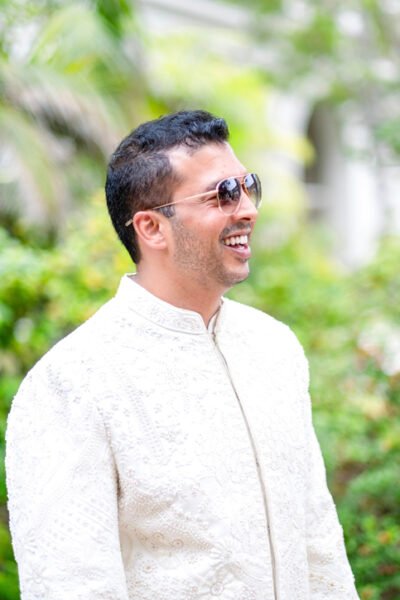Outdoor portrait of a South Asian groom in an ivory sherwani and sunglasses, smiling during his coastal wedding day.