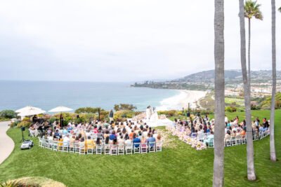 Wide view of a clifftop wedding ceremony overlooking the ocean with guests seated on the lawn and lush floral arrangements.