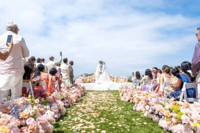 Bride walking down a flower-lined aisle toward the ceremony altar at a romantic outdoor coastal wedding.