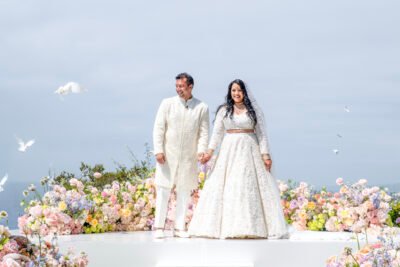 South Asian bride and groom holding hands on a white ceremony platform lined with pastel flowers at an elegant outdoor wedding.