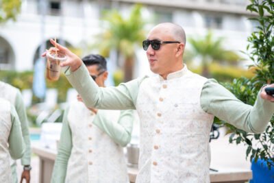 Groomsman in an ivory sherwani and sunglasses gesturing playfully during the outdoor wedding celebration.