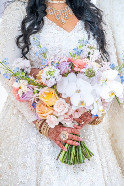 Closeup of South Asian bridal bouquet with pastel roses, orchids, and henna-adorned hands with bangles.