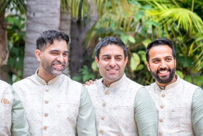 Group portrait of three groomsmen in coordinating ivory sherwanis standing in a garden.