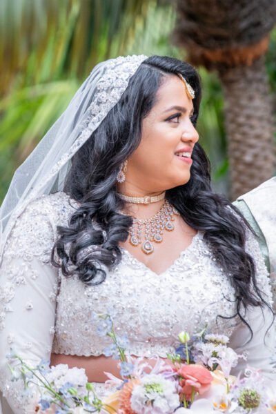 Closeup portrait of a South Asian bride in an ivory dress and veil, with long dark curls and layered jewelry, looking to the side.
