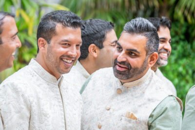 Closeup portrait of a smiling Indian groom in ivory sherwani in front of lush palm trees.