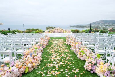 Lush floral aisle covered in petals leading to an oceanfront wedding altar with white chairs on either side.