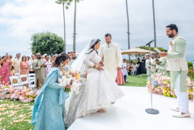 Bride and groom walking together down a white platform aisle lined with colorful flowers and family.