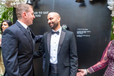 Groom in a tuxedo greeting a guest and smiling during a black-tie wedding reception.